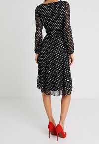 Robe noire à pois avec manches longues transparentes, taille cintrée et ourlet évasé. Des talons hauts rouges complètent l'esthétique contrastée de la tenue.