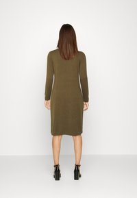 Robe longue vert olive à manches longues, coupe droite, texture côtelée et ourlet au genou. Chaussures noires à talons carrés avec des brides de cheville.
