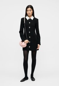 Γυναίκα που φοράει μαύρο φόρεμα με λευκό γιακά, μεγάλα διακοσμητικά κουμπιά, μαύρες καλτσοδέτες, μαύρες μπαλαρίνες και κρατάει ένα μικρό ροζ τσαντάκι.