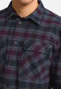 Chemise en flanelle à carreaux dans des tons de bordeaux, bleu marine et bleu clair. Elle dispose d'un col button-down, de deux poches poitrine et de boutons noirs. Tissu texturé.