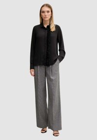 Camicia nera a maniche lunghe con dettagli di frange sul davanti, abbinata a pantaloni grigi a gamba larga con motivo e sandali neri.