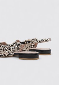 Sandalias tejidas en negro y crema con tira en el tobillo, que presentan una suela plana y un acabado liso y texturizado. Los detalles incluyen una hebilla dorada.