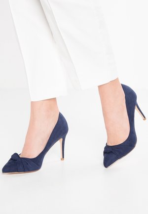 Granatowe zamszowe szpilki z detalem w postaci węzła na palcach, o spiczastym nosku i smukłym, stiletto obcasie.