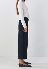 Kvinna klädd i en vit långärmad topp, svarta högmidjade vida byxor och svarta spetsiga flats, stående i profil mot en vit bakgrund.