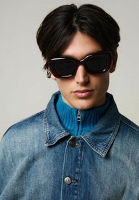 Denimjacka med två silverknappar ovanpå en ribbad blå polotröja. Modellen bär stora mörka solglasögon med en tjock burgunderram.