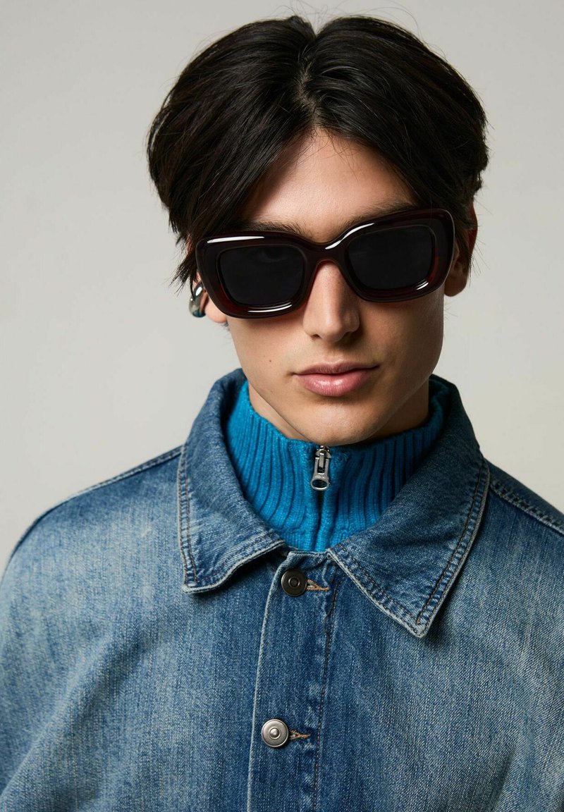 Denimjacka med två silverknappar ovanpå en ribbad blå polotröja. Modellen bär stora mörka solglasögon med en tjock burgunderram.