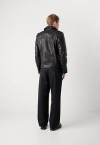 Man standing facing away, wearing a black leather jacket, loose black pants, and black shoes against a plain white background.