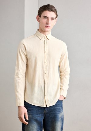 Chemise crème à boutons avec une texture douce, manches longues et col, associée à un jean bleu foncé. Design minimaliste, coupe décontractée.