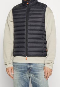 Mann trägt eine schwarze gesteppte Zip-Weste über einem beigefarbenen Langarmshirt und ausgebleichten blauen Jeans mit einem kleinen Riss in der Nähe der Tasche.