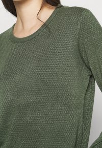 Grüner Strickpullover mit strukturiertem Muster, rundem Ausschnitt und langen Ärmeln. Der Stoff wirkt weich und leicht glänzend.