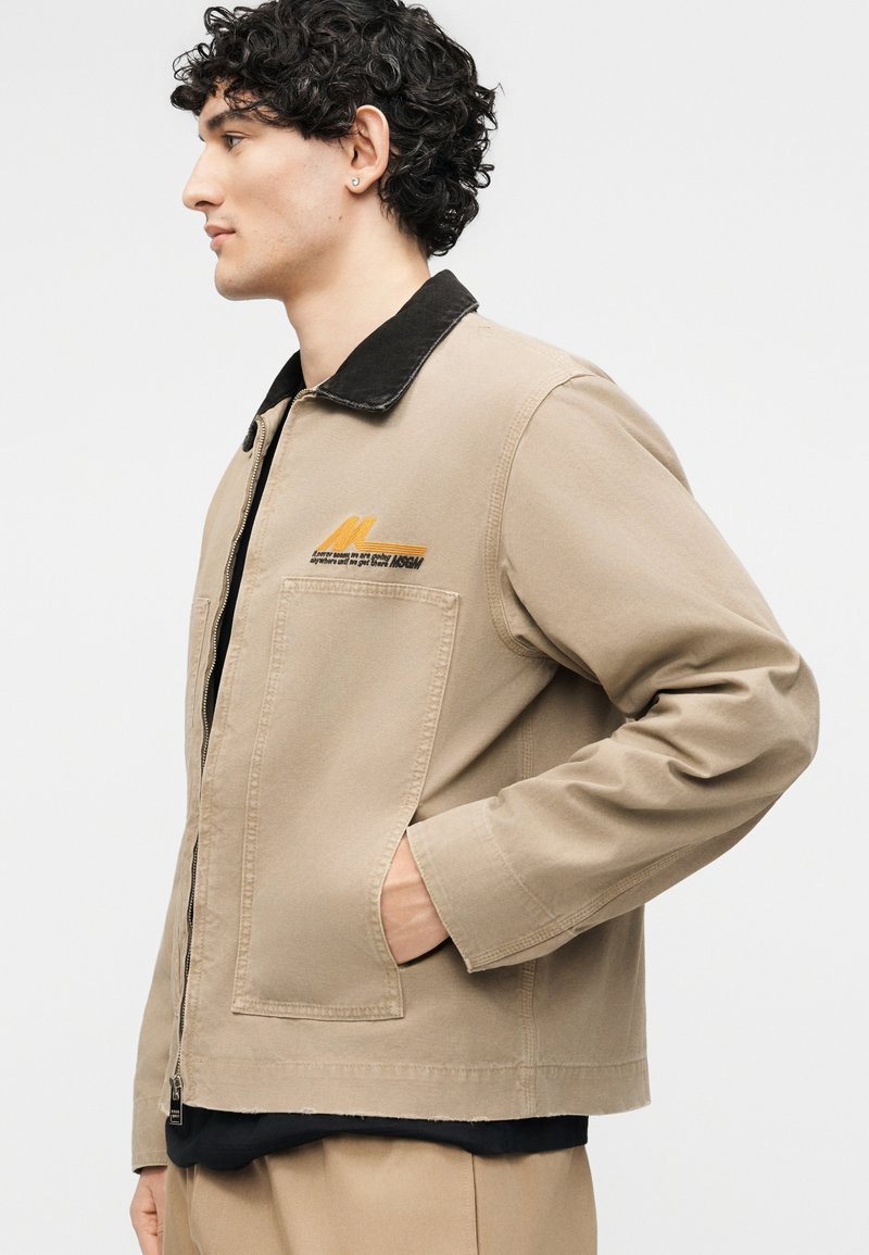 Jonge man met krullend haar, een beige jas met een zwarte kraag en een ritssluiting aan de voorkant, hand in de zak, kijkend naar links tegen een effen achtergrond.
