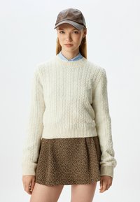 Crème kabelgebreide trui met geribbelde manchetten, gecombineerd met een bruin houndstooth rok. Het model draagt een bruine leren pet en een lichtblauw overhemd met kraag.