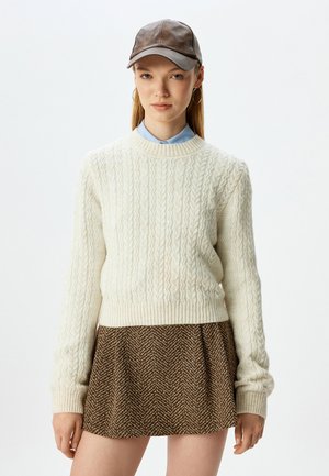 Crème kabelgebreide trui met geribbelde manchetten, gecombineerd met een bruin houndstooth rok. Het model draagt een bruine leren pet en een lichtblauw overhemd met kraag.