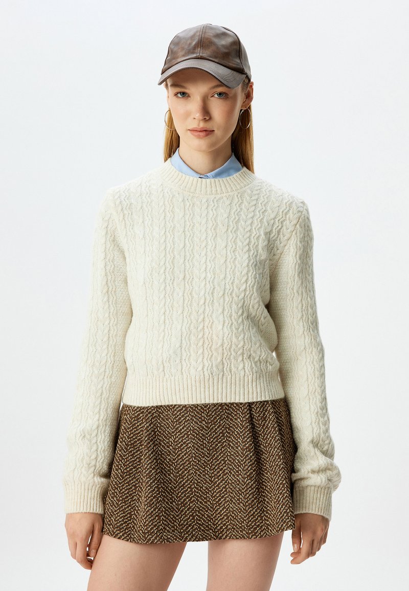Crème kabelgebreide trui met geribbelde manchetten, gecombineerd met een bruin houndstooth rok. Het model draagt een bruine leren pet en een lichtblauw overhemd met kraag.