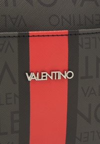 Texturované pozadí v černé a šedé barvě s vertikálním červeným pruhem a stříbrným logem "VALENTINO" umístěným na středu pruhu.