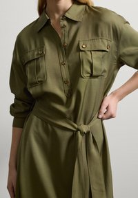 Olivengrünes, tailliertes Hemdkleid mit einem kragenlosen Ausschnitt, durchgehender Knopfleiste und zwei Brusttaschen. Glattes Material mit einer weichen Textur.