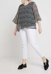 Femme portant un chemisier ample à pois noir et blanc, un pantalon blanc court et des mocassins à franges noirs, se tenant devant un fond uni.