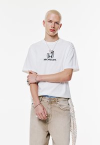 Hvid kortærmet t-shirt med et sort "HONDA" logo, parret med lysebeige denim jeans. Inkluderer et armbånd og et mønstret tørklæde om taljen.
