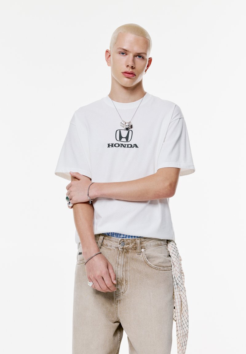Hvid kortærmet t-shirt med et sort "HONDA" logo, parret med lysebeige denim jeans. Inkluderer et armbånd og et mønstret tørklæde om taljen.