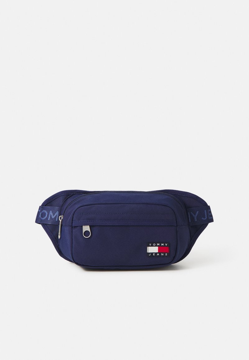 Navy fabric fanny pack with a zippered compartment, adjustable strap, and a logo patch featuring red, white, and blue colors. Textured surface.