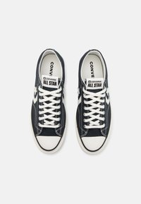 Tamno plave platnene tenisice s bijelim vezicama, gumenim vrhom i "CONVERSE ALL STAR" logotipom na jeziku i bočnoj strani. Sadrži bijele šavove.
