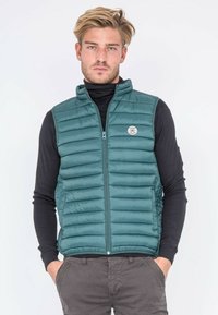 Gilet rembourré teal avec un col montant et une fermeture éclair à l'avant. Présente des coutures horizontales, un patch logo et des poches latérales. Porté sur un haut noir à manches longues.