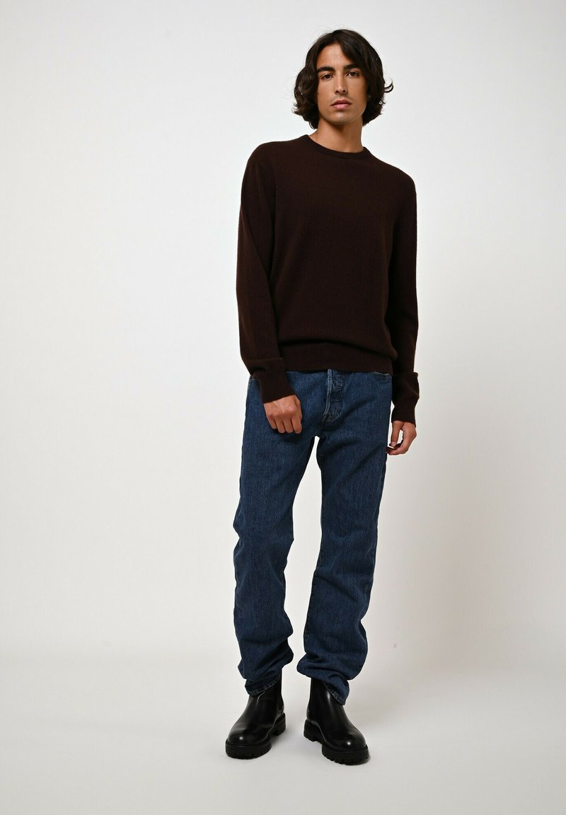Ciemnobrązowy sweter dzianinowy z okrągłym dekoltem, w zestawieniu z niebieskimi dżinsami. Na nogach czarne, masywne buty na grubej podeszwie. Prosty design.