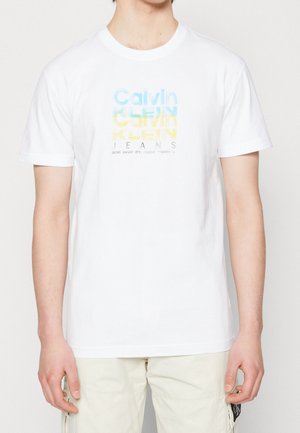 T-shirt en coton blanc avec un imprimé graphique comportant "Calvin Klein Jeans" en texte superposé bleu et jaune. Manches courtes, col rond.