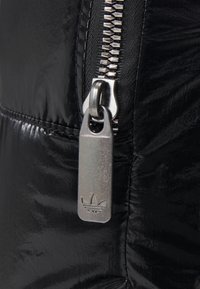 Svart puffigt tyg med en glänsande yta. Har en silverfärgad dragkedja med en märkesdragare i metall som visar en tredelad logotyp.