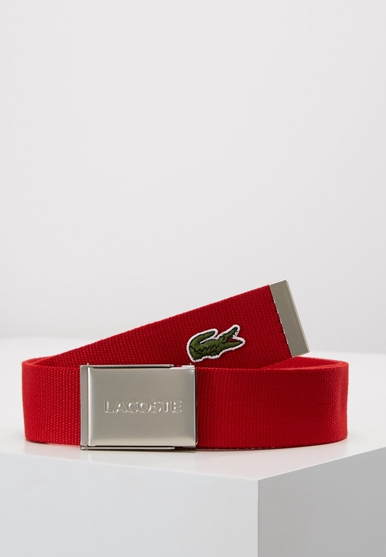 Ceinture en tissu rouge avec boucle métallique argentée gravée "Lacoste" et patch logo crocodile vert sur la sangle, présentée sur une surface blanche.