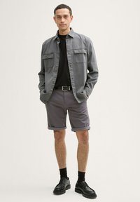 Camisa gris de botones con dos bolsillos en el pecho, camiseta negra debajo, pantalones cortos grises enrollados, calcetines negros y zapatos negros.