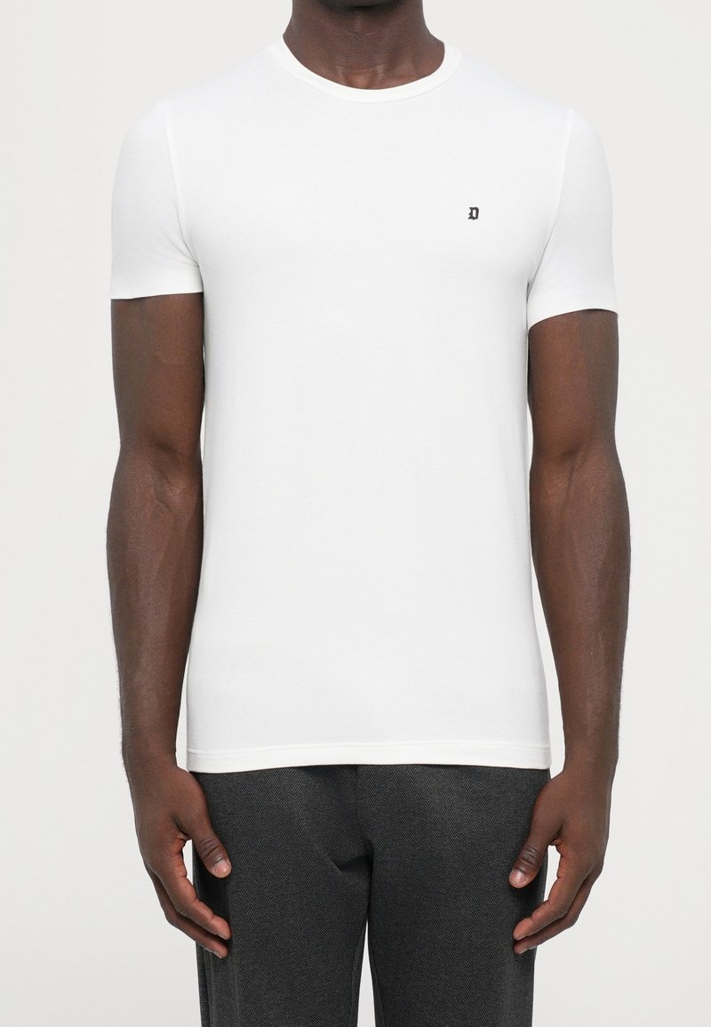Homme portant un t-shirt blanc uni cintré à manches courtes avec un petit logo noir "D" sur la poitrine et un pantalon gris foncé.