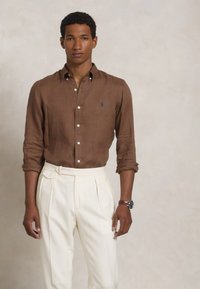 Kemeja linen coklat dengan kancing, celana putih berpita, dan sebuah jam tangan. Kemeja tersebut memiliki logo kecil, dan celana tersebut dilengkapi dengan pengencang di samping.