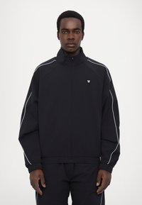 Svart zip-up jacka med vita pipingar, ribbat mönster, hög krage och logotyp på bröstet. Slätt tyg, avslappnad passform.