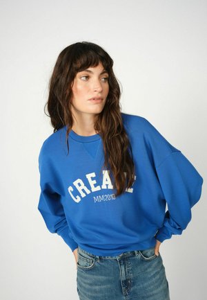 Blauwe katoenen sweatshirt met verlaagde schouders en een wit "CREATE" logo. Gecombineerd met lichtblauwe denim jeans, wat een casual ontwerp toont.