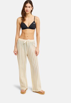 Młoda kobieta ubrana w czarny top bikini, luźne spodnie w beżowo-białe paski z sznurkiem oraz beżowe japonki na platformie, stojąca na białym tle.