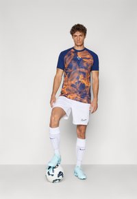 Short-sleeve, blue and orange patterned athletic shirt paired with white shorts and matching socks. Model poses with a soccer ball.