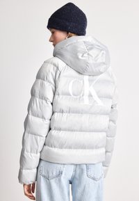 Svijetlosivi puffer s kapuljačom, s horizontalno ušivenim sekcijama i velikim bijelim logotipom na leđima. Nositi s plavim trapericama.