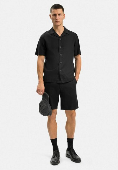 Svart kortärmad skjorta med krage och knappar, parat med svarta shorts. Modellen håller en svart keps, bär svarta skor och strumpor.