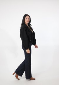 Sort cropped jakke med ballonærmer og guldknapper, kombineret med mørke, vidde jeans og brune højhælede støvler, sat mod en hvid baggrund.