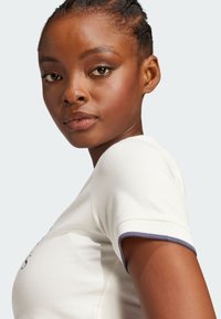 Pamuk majica u boji krem s tijesnim krojem, tamnoplavi obrub na rukavima, s tiskanim grafikama na prednjoj strani. Mekana tekstura i kratki rukavi.