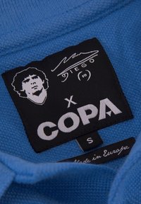 Blauwe stoffen kraag van een trui met een zwart label met witte tekst, een afbeelding van een gezicht en het "COPA" logo. Het maatlabel geeft "S" aan en "Made in Europe."