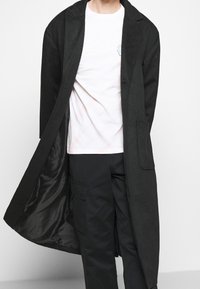 Persona che indossa un lungo cappotto nero sopra una t-shirt bianca e pantaloni cargo neri con uno sfondo chiaro uniforme.