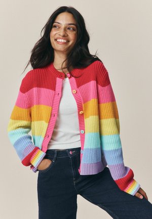 Woman smiling wearing multicolored striped knit cardigan over white shirt and dark jeans, standing with one hand in pocket against neutral background.