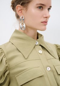 Blusa verde oliva con maniche a palloncino, grande colletto e due tasche sul petto, con bottoni in perla. Il modello indossa orecchini ovali d'argento e oro.
