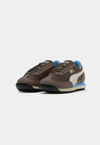 Ein Paar braune Puma Easy Rider Sneaker mit weißen Seitenstreifen und blauer Innenauskleidung, schräg positioniert, um Vorder- und Seitenansicht auf weißem Hintergrund zu zeigen.