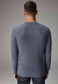 Uomo con capelli corti castani, vestito con un maglione grigio lavorato a maglia e jeans scuri, visto di spalle su uno sfondo semplice.