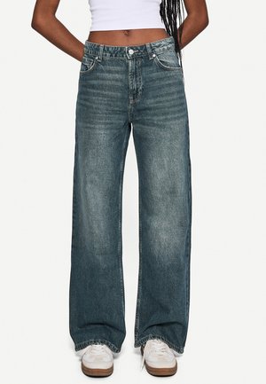 Person iført højttaljede, vide, udvaskede blå jeans med hvide sneakers og en kort hvid top, stående mod en ensfarvet baggrund.