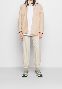 Beige cordjacka med två framfickor, lager-på-lager över en vit hoodie. Krämfärgade tapered joggers och gröna, mönstrade sneakers kompletterar stilen.