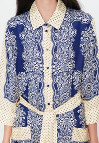 Gros plan d'une robe bleue et crème avec des motifs floraux complexes et de petits pois, dotée d'une ceinture nouée à la taille et d'une patte de boutonnage à l'avant.